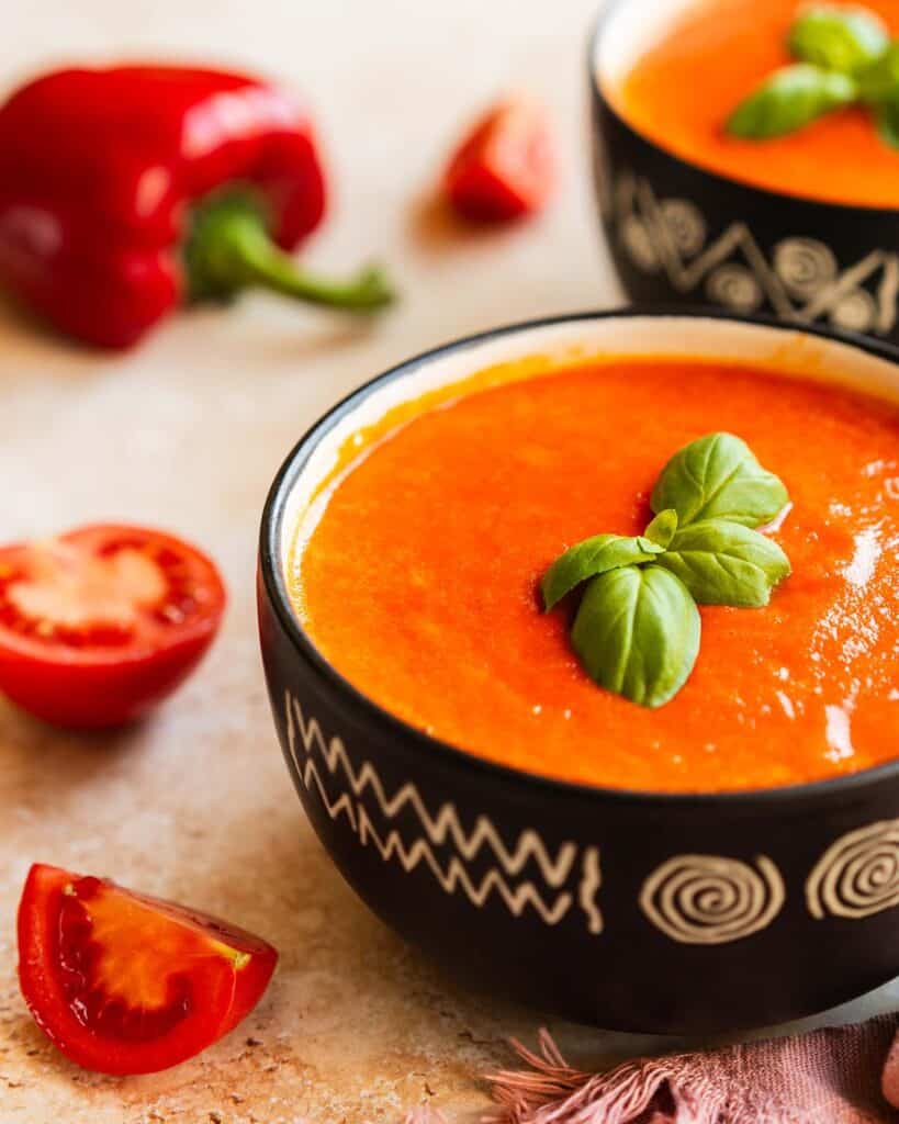 A bowl of roasted red pepper and tomato soup, garnished with fresh basil leaves. Image by Jaz Kitchen